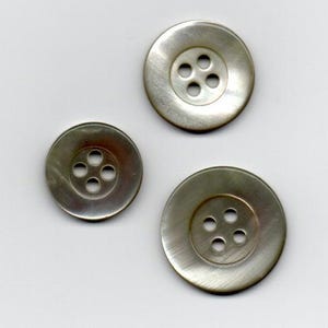 Peut inclure: Trois boutons ronds gris irisés avec quatre trous chacun. Les boutons varient en taille, avec une zone centrale légèrement surélevée contenant les trous. Les boutons sont posés sur un fond blanc uni.
