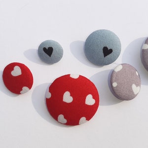 May include: Assortment of fabric covered buttons with different patterns. The buttons are red with white hearts, gray with white hearts, and blue with black hearts. The text "Euro-Band" is visible in the image.