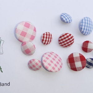 Puede incluir: Surtido de botones cubiertos de tela en varios colores y patrones, incluyendo vichy rosa y blanco, vichy rojo y blanco, vichy azul y blanco y vichy azul marino y blanco. Los botones están dispuestos sobre una superficie blanca. Euro-Band.
