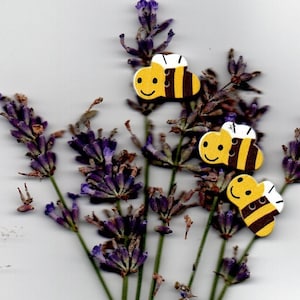 Może przedstawiać: Trzy drewniane guziki w kształcie pszczoły z żółto-czarnymi paskami i białymi skrzydłami. Guziki znajdują się na tle fioletowych kwiatów lawendy.