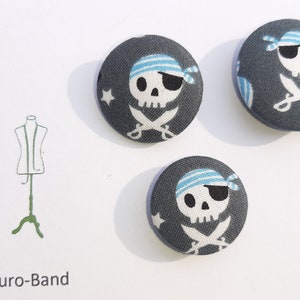 May include: Three gray fabric buttons with a white skull and crossbones design. The skull is wearing a blue and white striped eye patch and a blue and white striped bandana. The buttons are approximately 1 inch in diameter.