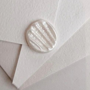 Puede incluir: Primer plano de un sobre de papel blanco texturizado con un sello de cera nacarado. El sello es redondo con un diseño estriado. El papel tiene una textura sutil y orgánica.