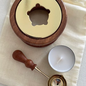 Pode incluir: Um kit de selagem de cera com uma base de madeira e uma inserção de metal dourado com um recorte decorativo. Inclui uma pequena colher de metal com cabo de madeira e uma vela de chá branca. O kit repousa sobre um tecido bege.