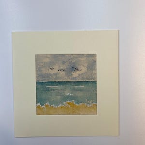 May include: A square watercolour painting of a coastal scene featuring a yellow sandy beach, turquoise ocean waves with white foam, and a cloudy blue sky with three birds flying in the distance, all set within a cream-coloured paper mat border.