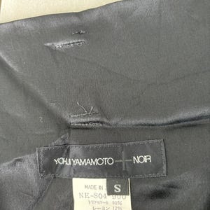 May include: Black garment with a label that reads "YOHJI YAMAMOTO + NOIR." The label also includes the text "MADE IN" and "S." The garment's fabric composition is listed on the label.