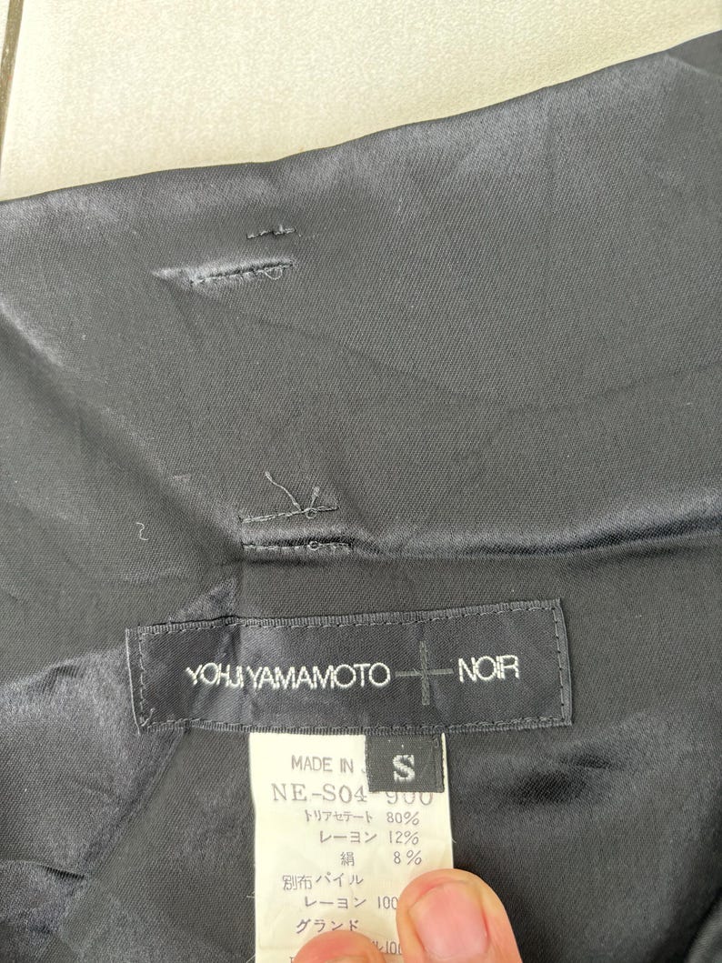 May include: Black garment with a label that reads "YOHJI YAMAMOTO + NOIR." The label also includes the text "MADE IN" and "S." The garment's fabric composition is listed on the label.