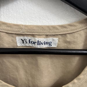 以下が含まれることがあります： ベージュ色の衣服のクローズアップ。白のラベルには紺色の文字で「Y's for living」と書かれています。衣服は黒いハンガーに掛けられ、白い壁を背景にしています。