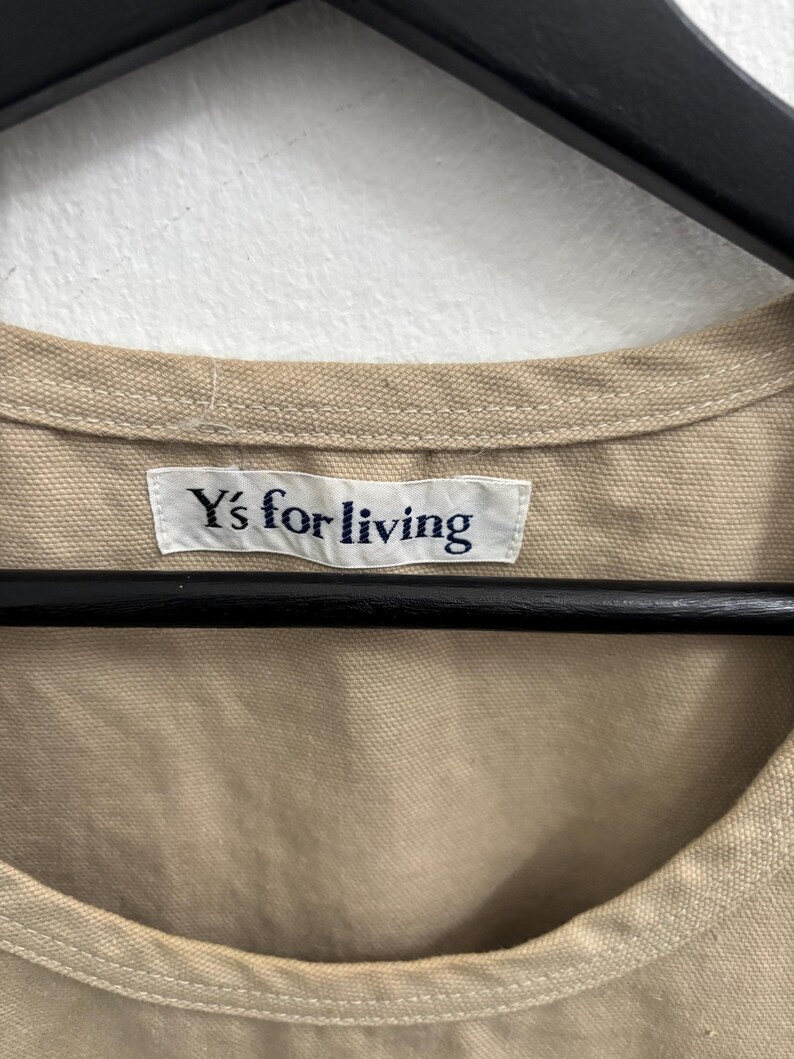 以下が含まれることがあります： ベージュ色の衣服のクローズアップ。白のラベルには紺色の文字で「Y's for living」と書かれています。衣服は黒いハンガーに掛けられ、白い壁を背景にしています。