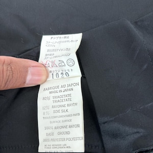 May include: A close-up of a black garment with a white tag. The tag has text in Japanese and French, including fabric composition: 80% Triacetate, 12% Rayonne Rayon, and 8% Silk. The garment is made in Japan.