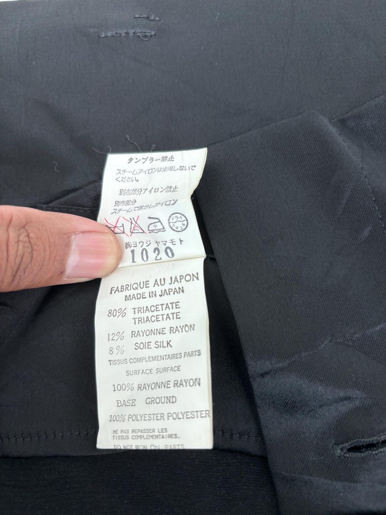 May include: A close-up of a black garment with a white tag. The tag has text in Japanese and French, including fabric composition: 80% Triacetate, 12% Rayonne Rayon, and 8% Silk. The garment is made in Japan.