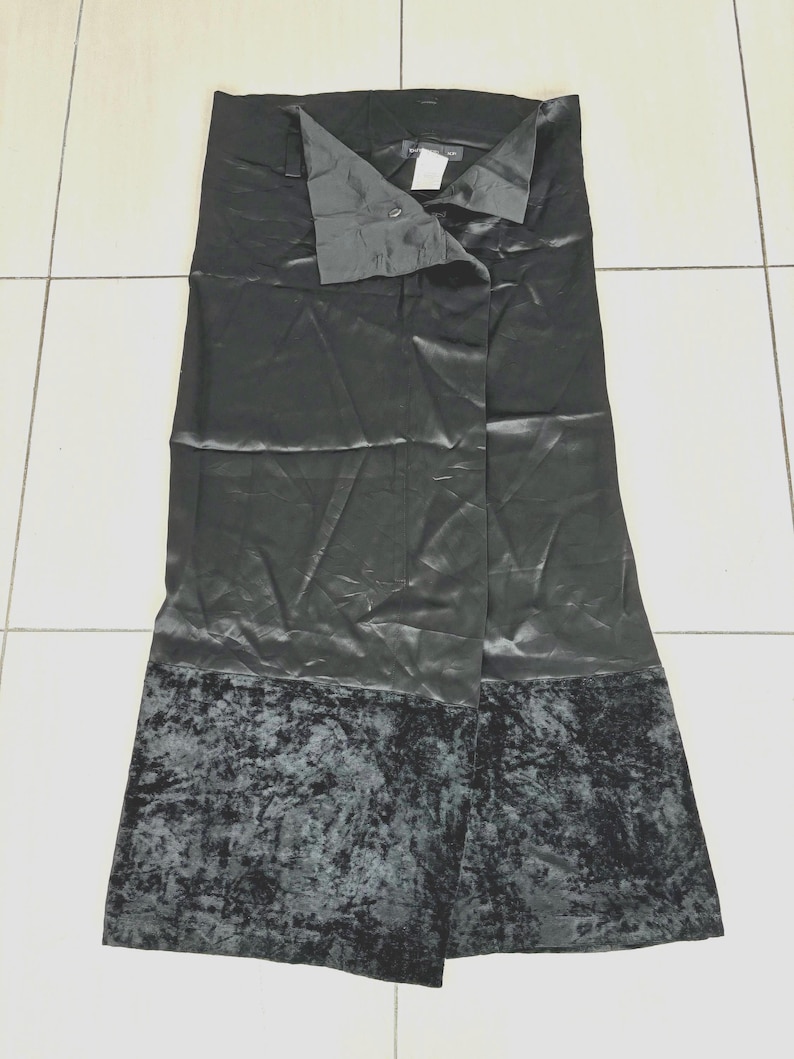 May include: A black skirt with a satin finish and a velvet hem. The skirt has a button closure at the waist and a split front design. The skirt is a midi length.