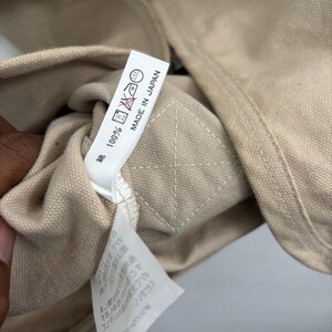 以下が含まれることがあります： ベージュ色の衣類タグのクローズアップ。テキスト「100%」と「MADE IN JAPAN」が記載されています。タグにはケアマークも表示されています。衣類は明るい色の生地で作られているようです。