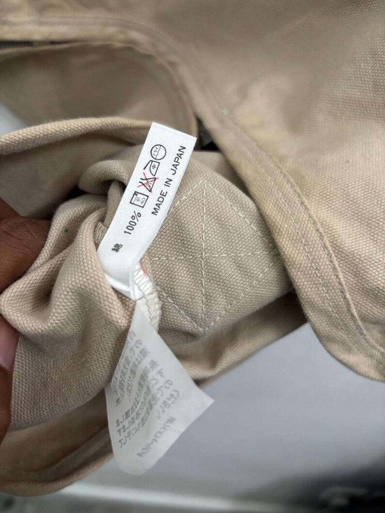 以下が含まれることがあります： ベージュ色の衣類タグのクローズアップ。テキスト「100%」と「MADE IN JAPAN」が記載されています。タグにはケアマークも表示されています。衣類は明るい色の生地で作られているようです。