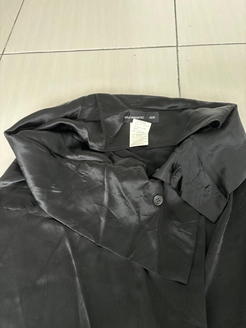 May include: Black satin top with a unique asymmetrical collar and a single button closure. The top has a smooth, shiny texture and a relaxed fit. The tag reads "YOHJI YAMAMOTO NOR".