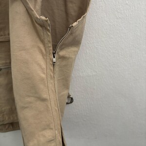 以下が含まれることがあります： 前面に銀色のジッパーが付いた、ベージュ色のキャンバスベストのクローズアップ。ベストには複数のポケットと金属リングのディテールがあります。生地は丈夫で耐久性があり、アウトドアでの使用に適しています。