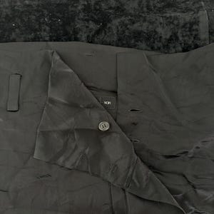 May include: Black garment with a shiny, textured appearance. The garment has a button closure and a folded collar. The word "NOR" is visible on a label. The top portion of the image shows a dark, textured fabric.
