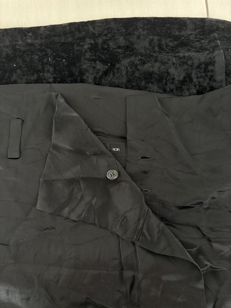 May include: Black garment with a shiny, textured appearance. The garment has a button closure and a folded collar. The word "NOR" is visible on a label. The top portion of the image shows a dark, textured fabric.