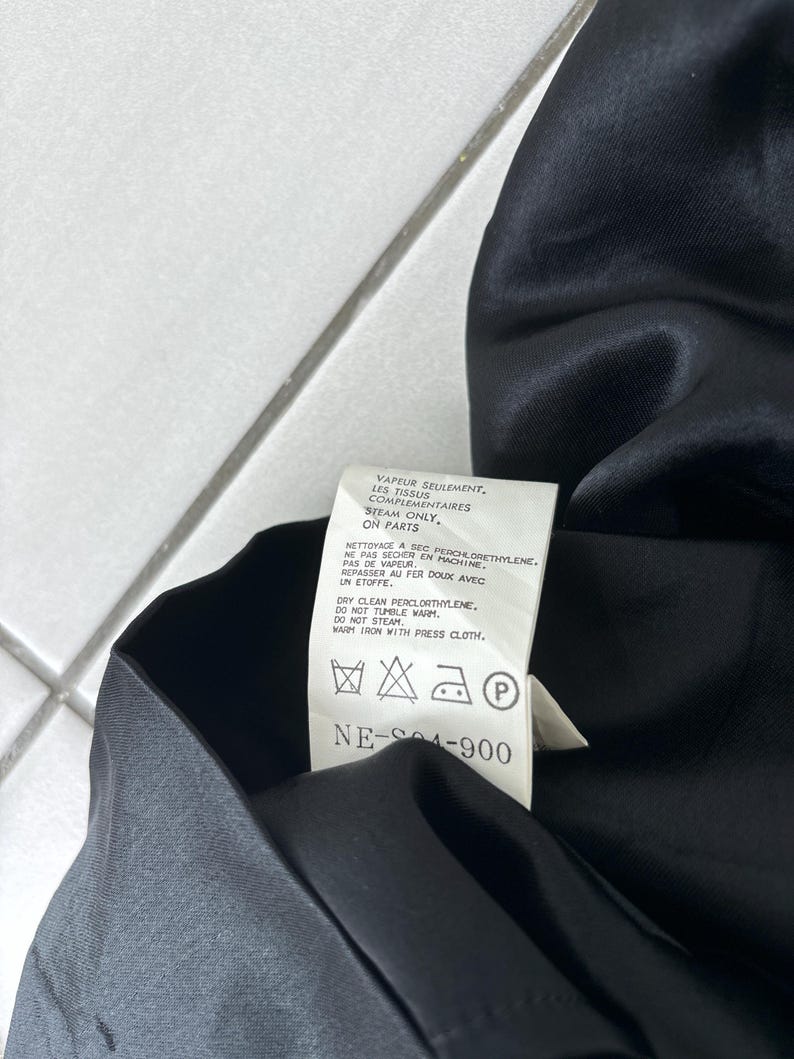May include: A close-up of a black garment with a care label attached. The label provides instructions for cleaning, including dry cleaning with perchloroethylene and warm ironing with a press cloth. The label also includes the text "NE-S04-900."