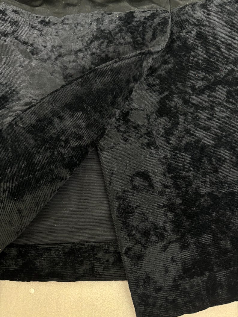 May include: Close-up of a black velvet fabric with a ribbed texture. The fabric is folded, revealing the underside and a contrasting smooth lining. The image highlights the luxurious texture and depth of the material.