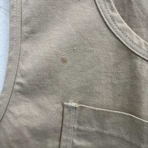 以下が含まれることがあります： ポケット付きのベージュ色のキャンバスエプロンのクローズアップ。エプロンにはステッチと小さな茶色のシミが見えます。生地は使い古されたように見えます。画像は作業服の質感と細部に焦点を当てています。