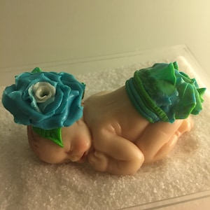 Könnte beinhalten: Eine schlafende Babyfigur aus Fondant, verziert mit einer blauen Rose und einer grün-blauen Windel. Die Figur liegt auf einer weißen Oberfläche.