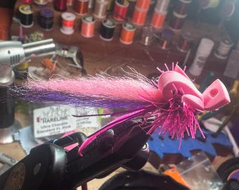 May include: A pink and purple fly tying material with a pink foam head. The fly is tied on a vise.