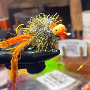 May include: A close-up of a fly fishing lure with orange and yellow feathers, a green and silver body, and a bright orange and yellow head.