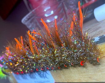 May include: A close-up of a group of fly fishing lures with orange and brown feathers and iridescent accents. The lures have black eyes and are arranged in a row on a wooden surface.