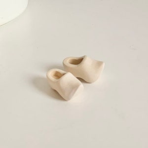 Könnte beinhalten: Ein Paar kleine, hellfarbene Holzschuhe liegen auf einer weißen Oberfläche. Ein weißer Pflanztopf mit braunem Rand und eine grüne Pflanze befinden sich im Hintergrund. Die Holzschuhe sind eine Miniaturversion traditioneller niederländischer Schuhe.