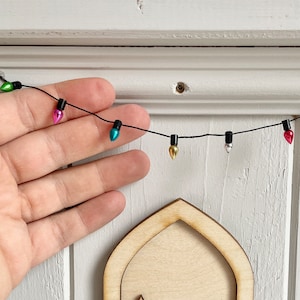Könnte beinhalten: Eine Miniatur-Feentür aus Holz mit einer kleinen Leiter und einer Lichterkette mit bunten Weihnachtslichtern. Die Tür ist hellbraun mit dekorativen Scharnieren. Die Lichter sind in verschiedenen Farben, darunter Grün, Rosa und Blau.