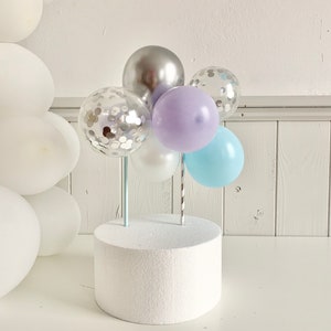 May include: A white cake stand with a cluster of balloons on top. The balloons are silver, light blue, lavender, and clear with silver confetti.