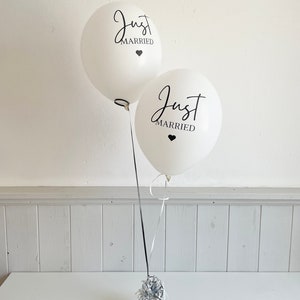 Peut inclure: Deux ballons blancs avec l'inscription noire "Just Married" et un petit c&oelig;ur noir. Les ballons sont attach&eacute;s ensemble avec un ruban noir et blanc et sont pos&eacute;s sur une table blanche.