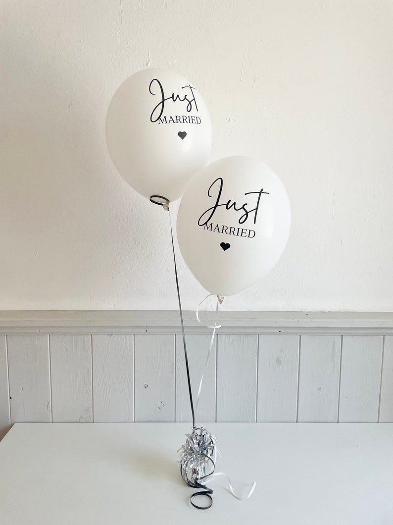 Peut inclure: Deux ballons blancs avec l'inscription noire "Just Married" et un petit c&oelig;ur noir. Les ballons sont attach&eacute;s ensemble avec un ruban noir et blanc et sont pos&eacute;s sur une table blanche.