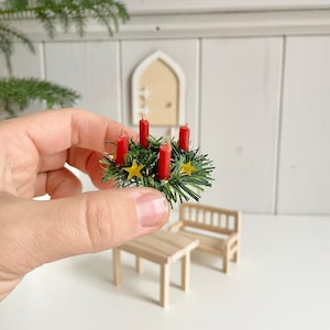 May include: Miniature Christmas wreath with four red candles, green foliage, and gold star accents. The wreath is held in a hand, with a small wooden table and bench in the background. A festive holiday decoration.