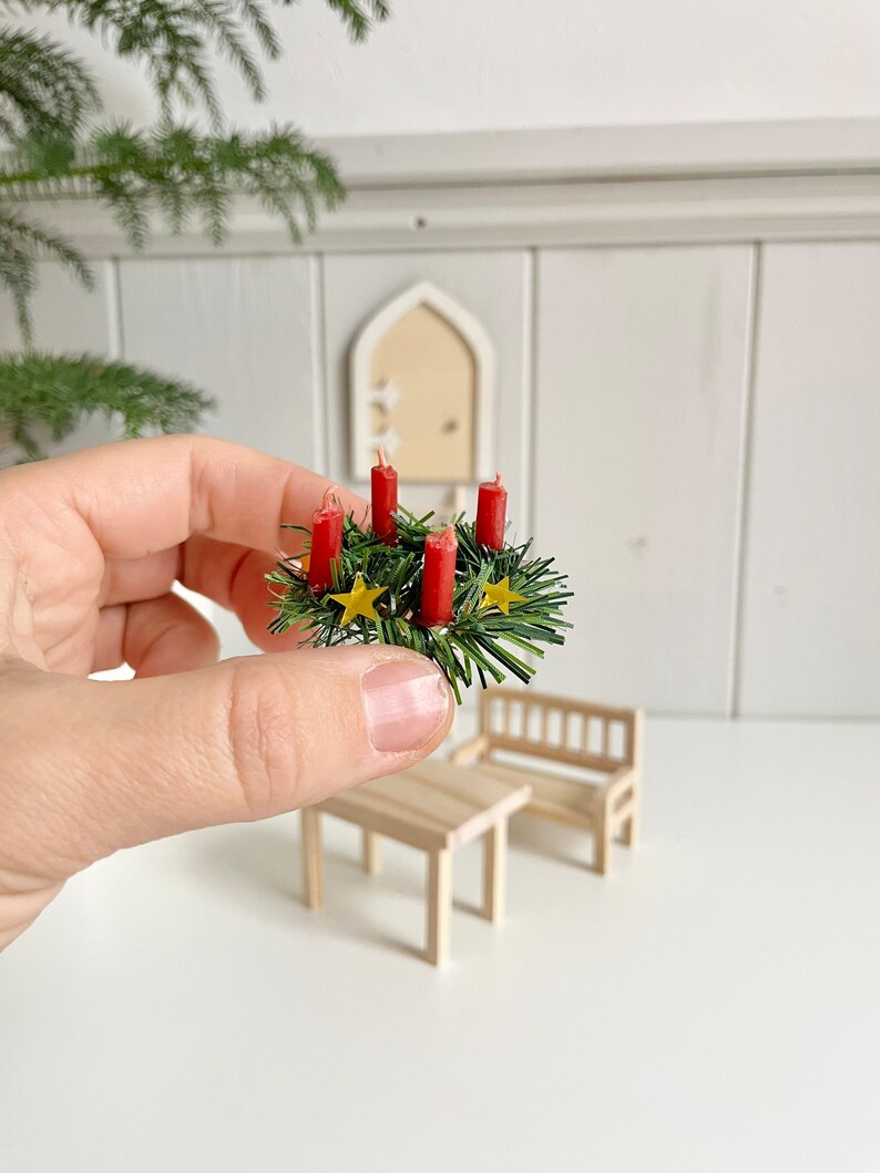 May include: Miniature Christmas wreath with four red candles, green foliage, and gold star accents. The wreath is held in a hand, with a small wooden table and bench in the background. A festive holiday decoration.