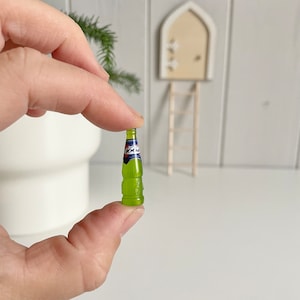 May include: A miniature green bottle with a blue and red label is held between fingers. The bottle has a textured design and a small, detailed label. The background includes a miniature door, ladder, and greenery, suggesting a fairy garden theme.