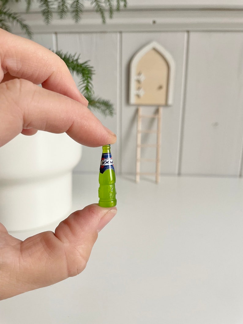 May include: A miniature green bottle with a blue and red label is held between fingers. The bottle has a textured design and a small, detailed label. The background includes a miniature door, ladder, and greenery, suggesting a fairy garden theme.