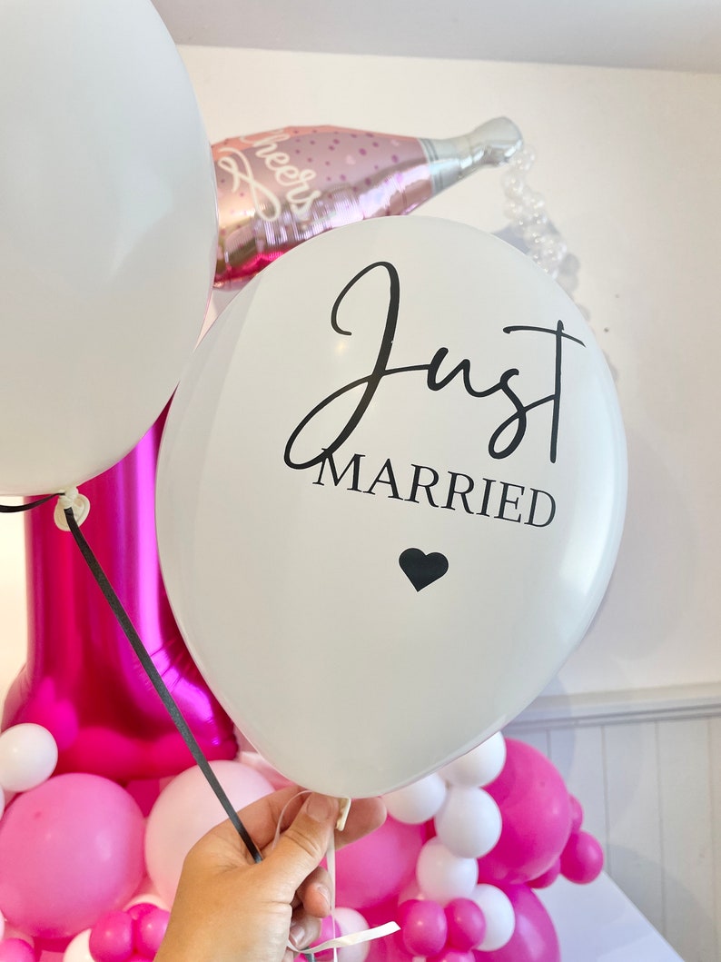 Peut inclure: Ballon blanc avec le texte "Just Married" en &eacute;criture noire avec un petit c&oelig;ur noir sous le texte. Le ballon est attach&eacute; avec un ruban blanc et est entour&eacute; de ballons roses et blancs.