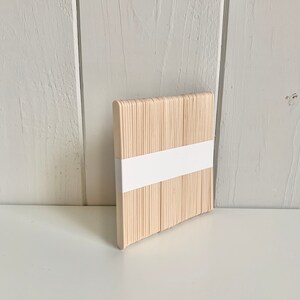 May include: A bundle of natural wooden craft sticks, approximately 100 sticks, arranged in a square shape. The sticks are bundled together with a white paper band.