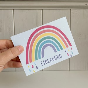 May include: A white card with a rainbow and raindrops. The text "EINLADUNG" is printed on the card.