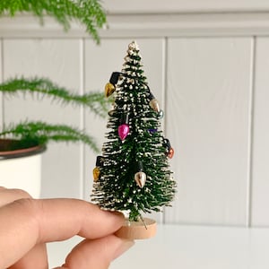 Könnte beinhalten: Ein kleiner Weihnachtsbaum mit grünen Nadeln und bunten Lichterketten. Der Baum ist mit kleinen goldenen, silbernen und rosa Glühbirnen geschmückt. Er steht auf einem kleinen, runden Holzsockel, ideal für festliche Dekorationen.
