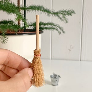 Könnte beinhalten: Miniatur-Besen mit einem Kopf aus Naturstroh und einem Holzgriff, mit rotem Faden gebunden. Ein kleiner, silberner Metalleimer steht daneben. Die Artikel sind wahrscheinlich für ein Puppenhaus oder ein Miniaturprojekt.