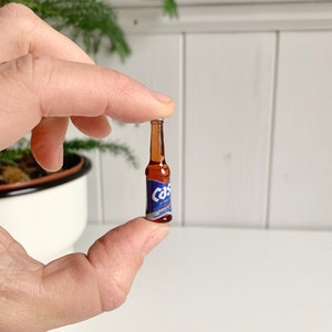May include: A miniature beer bottle with a brown glass body and a blue and white label. The label reads "Cas" in a stylized font. The bottle is held between two fingers against a white background.