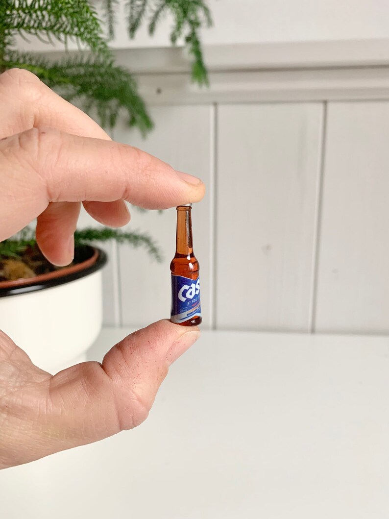 May include: A miniature beer bottle with a brown glass body and a blue and white label. The label reads "Cas" in a stylized font. The bottle is held between two fingers against a white background.