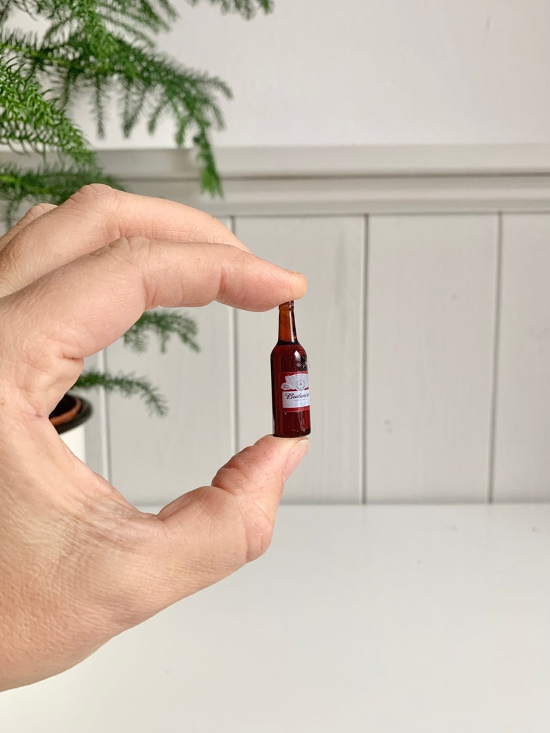 May include: Miniature brown glass bottle with a white and red label that reads "Budweiser". The bottle is held between fingers against a white background. The bottle is a small replica of a beer bottle.