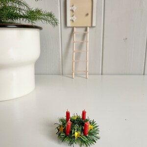 May include: A miniature Christmas wreath with four red candles and gold star accents. The wreath is surrounded by green faux pine needles. A small wooden door with a ladder is in the background, along with a white pot with a green plant.