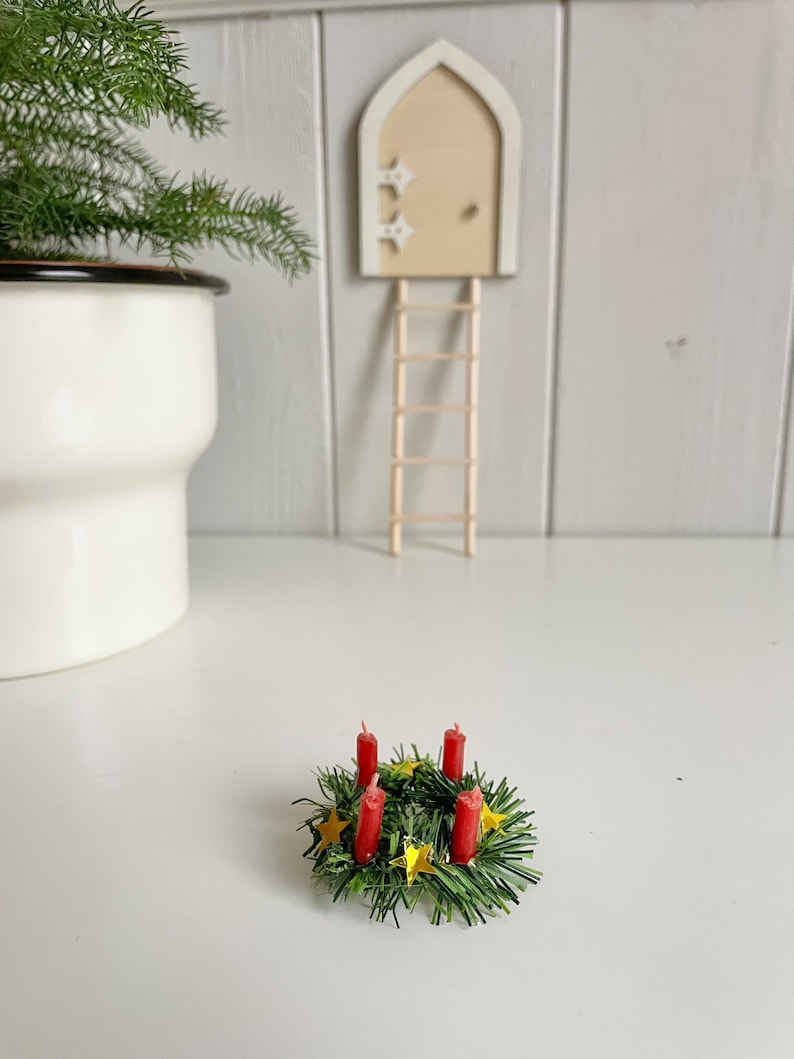 May include: A miniature Christmas wreath with four red candles and gold star accents. The wreath is surrounded by green faux pine needles. A small wooden door with a ladder is in the background, along with a white pot with a green plant.