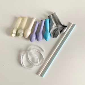 May include: A set of party supplies including balloons, straws, and ribbon. The balloons are in shades of blue, white, and gray. The straws are white and blue. The ribbon is white.