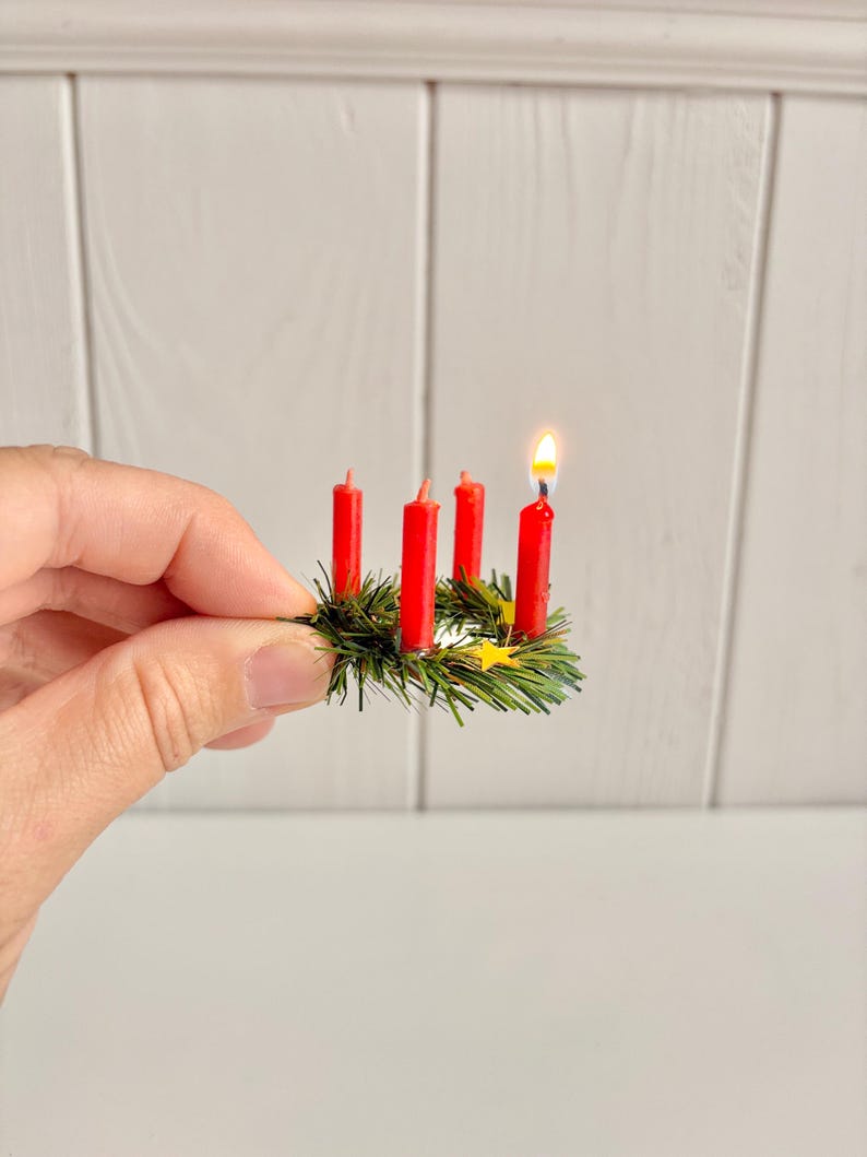 May include: Miniature Advent wreath with four red candles, one lit with a small flame. The candles are set in a green wreath with small yellow star accents. The wreath is held in a hand against a white background.