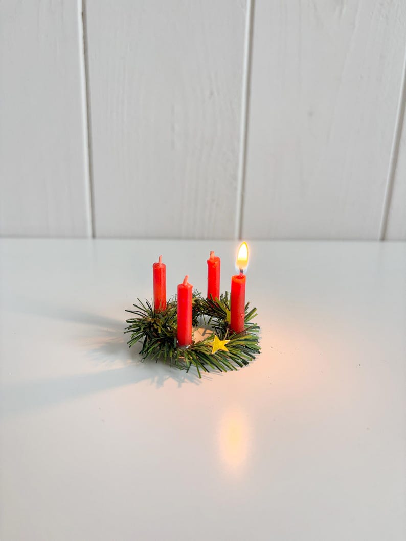 May include: A miniature Advent wreath with four red candles, one lit with a bright flame. The wreath is made of green faux pine needles and includes a small yellow star. The arrangement sits on a white surface.